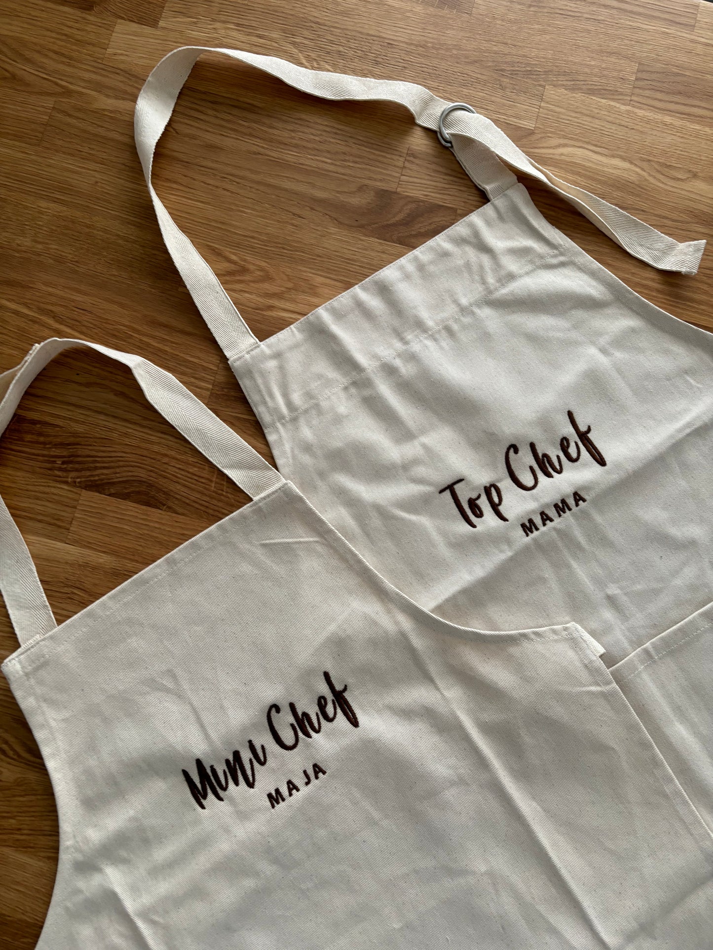 Zwei Schürzen jeweils mit braunem Garn bestickt. Die Junior Schürze ist mit Mini Chef Maja bestickt und die Erwachsenen Schürze mit Top Chef Mama.