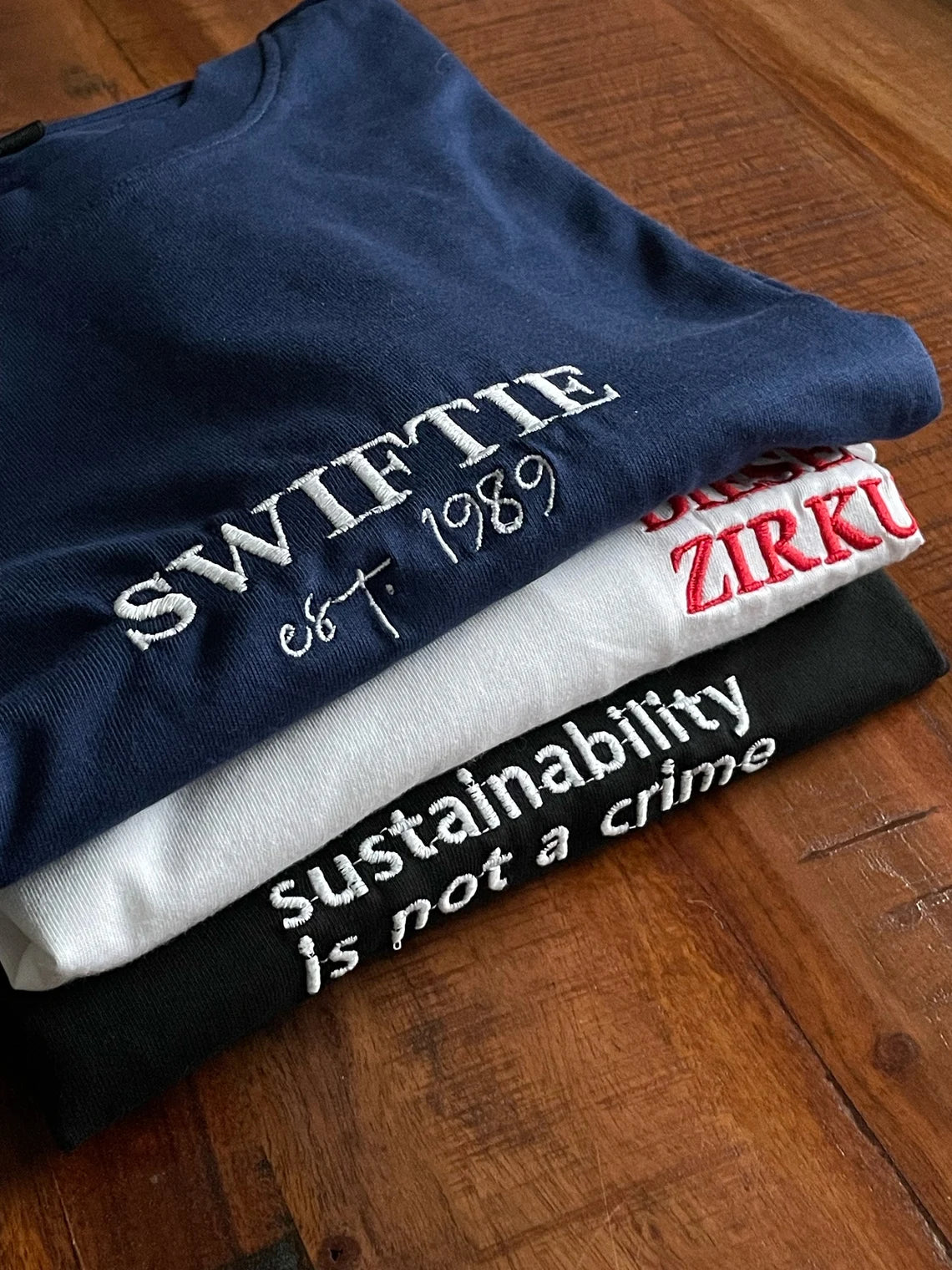 Drei T-Shirts mit jeweils einer personalisierten Bestickung. Das erste ist Navy Blau mit Swiftie. Ein weißes T-Shirt mit einer roten Bestickung und ein schwarzes mit weißem Stick.
