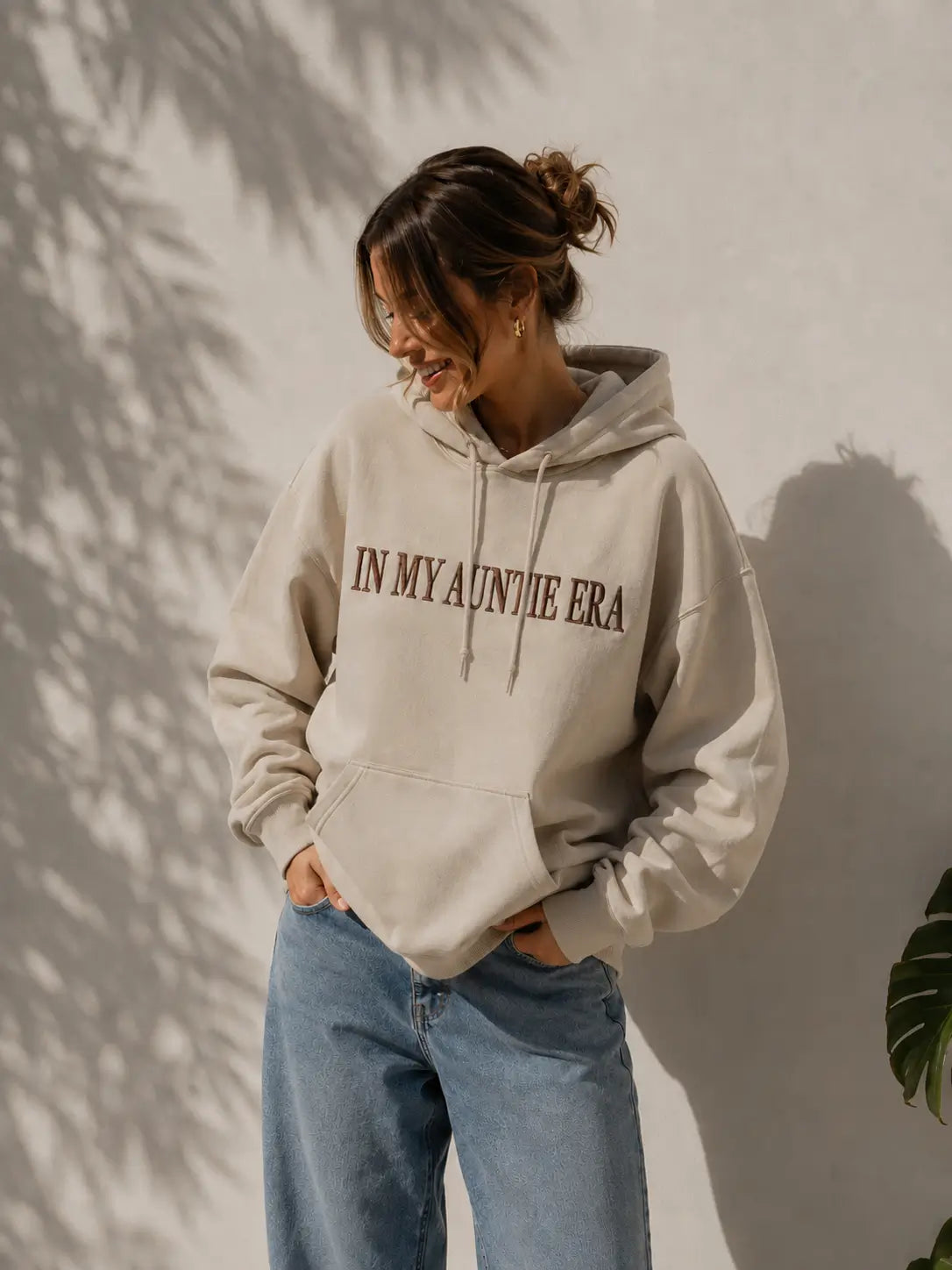 Eine Frau trägt einen personalisierten beigen Hoodie mit der Stickerei vorne "IN MY AUNTIE ERA".