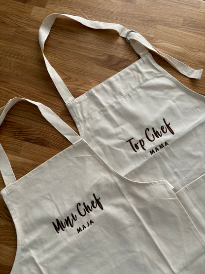 Zwei Schürzen jeweils mit braunem Garn bestickt. Die Junior Schürze ist mit Mini Chef Maja bestickt und die Erwachsenen Schürze mit Top Chef Mama.