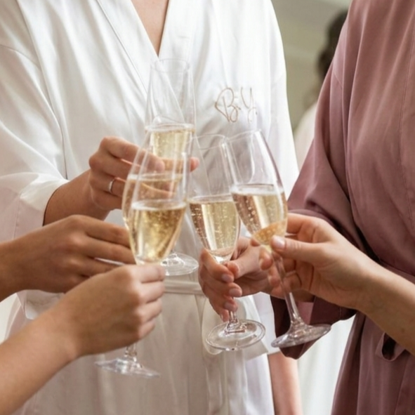 Die Braut und die Brautjungfern stoßen beim Getting Ready zusammen an. Die Braut trägt einen personalisierten Morgenmantel.