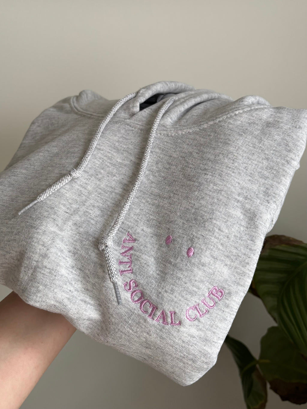 Ein personalisierter bestickter Hoodie in der Farbe Ash Heather mit einem Smiley "Anti Social Club" in lila.