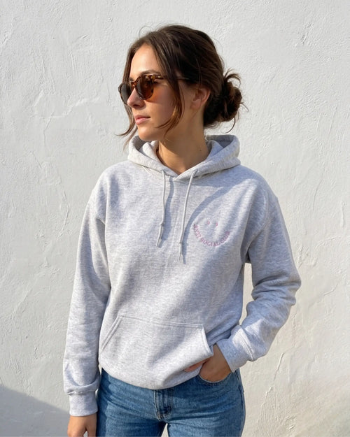 Eine Frau trägt einen Hoodie mit einem gestickten Smiley "Anti Social Club" in lila. 