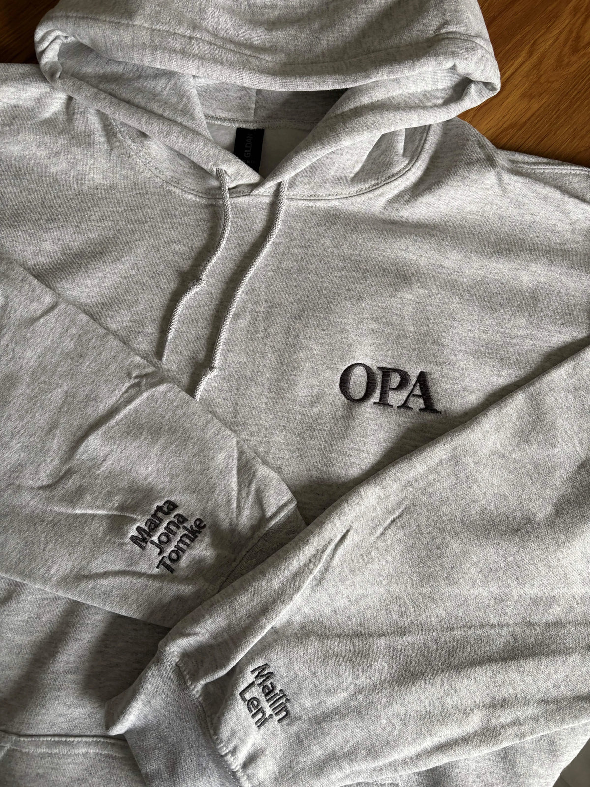 Ein Hoodie in der Farbe Ash Heather mit einer Stickerei in der Farbe Dark Grey: OPA. Auf dem rechten Ärmel sind drei Kindernamen gestickt: Marta, Jona und Tomke. Auf dem linken Ärmel sind zwei Kindernamen gestickt: Mailin und Leni.