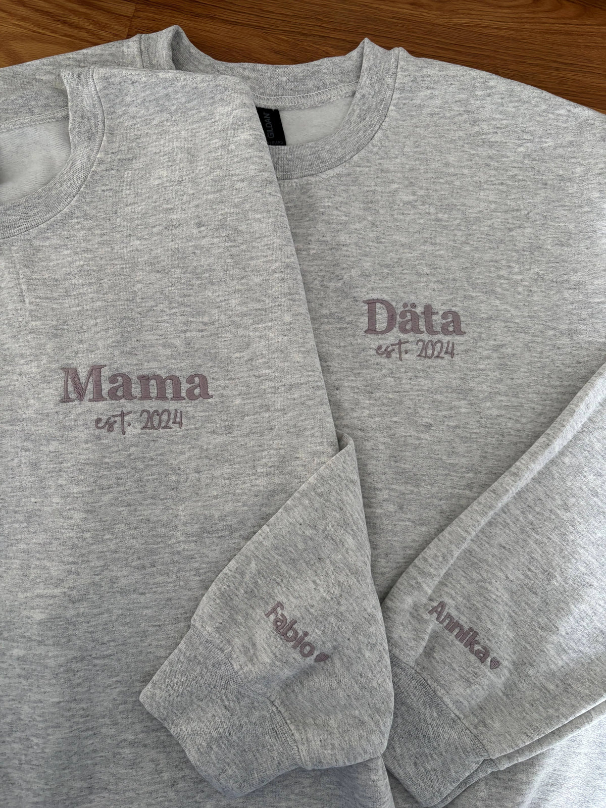 Zwei Sweater in der Farbe Ash Heather mit jeweils einer Stickerei in der Farbe Grau: Mama & Däta est. 2024. Es ist jeweils ein Kindername auf dem linken Ärmel: Fabio & Annika.