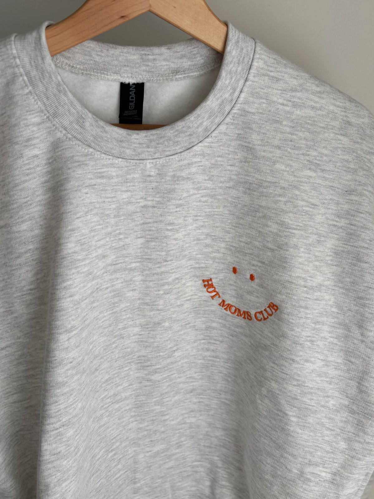 Ein grauer Sweater ist mit einem orangenen Smiley auf der Brust bestickt: Hot Moms Club.