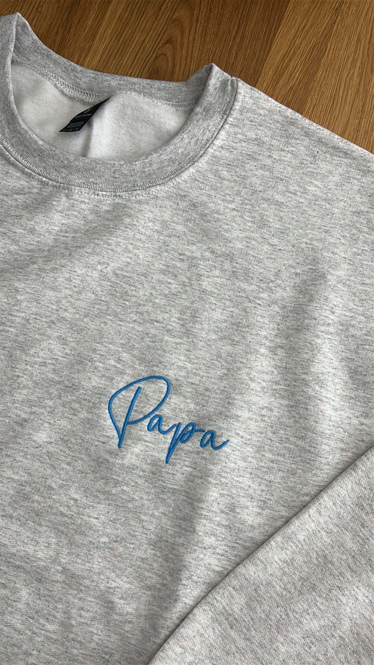 Ein Sweater in der Farbe Ash Heather mit einer Stickerei in der Farbe Aqua: Papa.