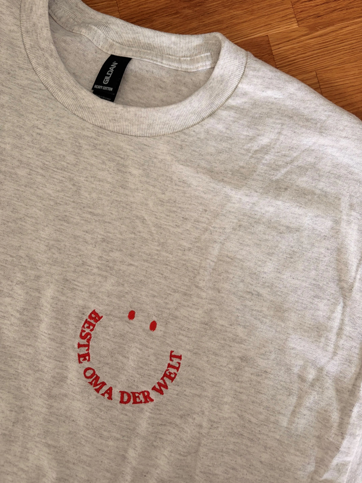 Ein T-Shirt in der Farbe Ash Heather mit kurzen Ärmeln und einen roten Smiley auf der linken Brustseite. Die Grafik besteht aus zwei kleinen ovalen Augen und einem geschwungenen Schriftzug BESTE OMA DER WELT, der ein lächelndes Gesicht bildet.