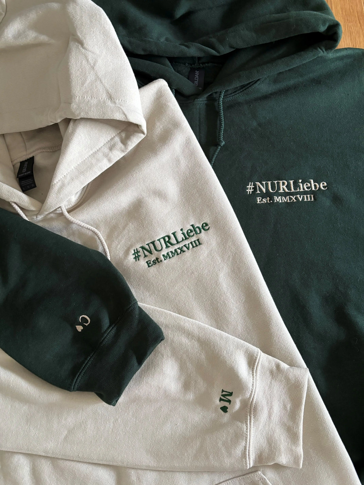 Zwei Hoodies in der Farbe Beige und Forest Green mit jeweils einer Stickerei links auf der Brust und auf dem Ärmel.