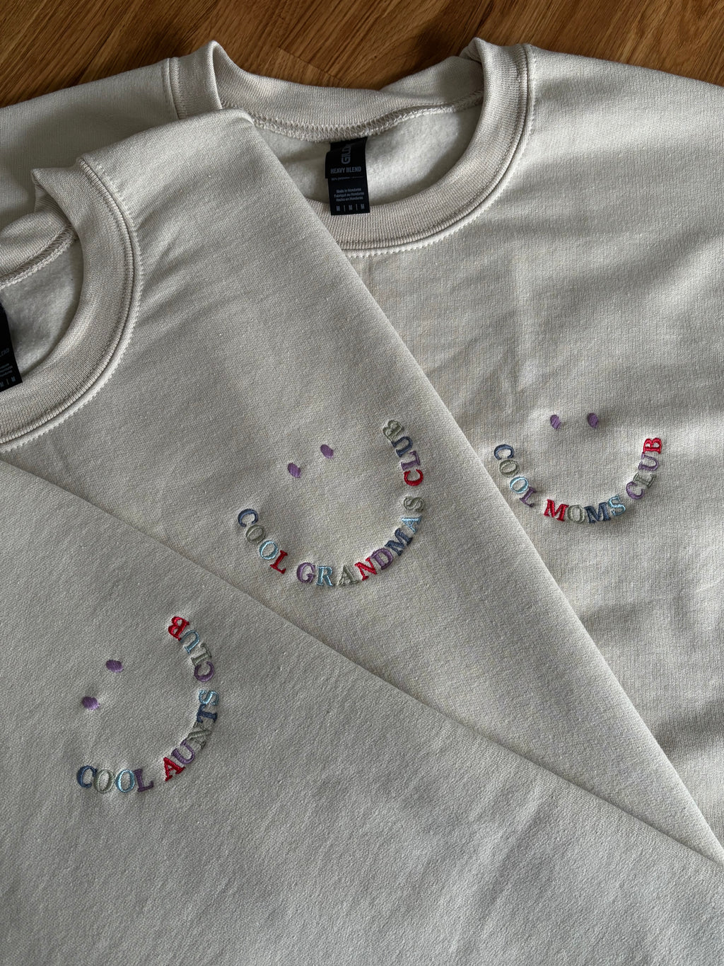 Hier sind drei Sweater, die farblich aufeinander abgestimmt sind. Alle drei haben eine Stickerei in Form eines Smileys mit buntem Garn: Cool Moms Club, Cool Moms Club und Cool Aunts Club.