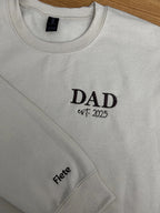 Ein beiger Sweater. Der Stick ist in dunkelbraun. Gestickt ist in Großbuchstaben DAD und darunter est. 2025. Auf dem Ärmel steht ein Kindername: Fiete.