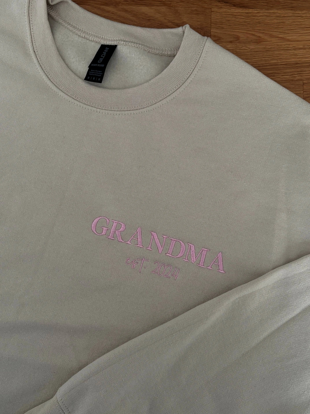 Ein Sweater in der Farbe Beige mit einer Stickerei in der Farbe Rosa: Grandma est. 2024. 