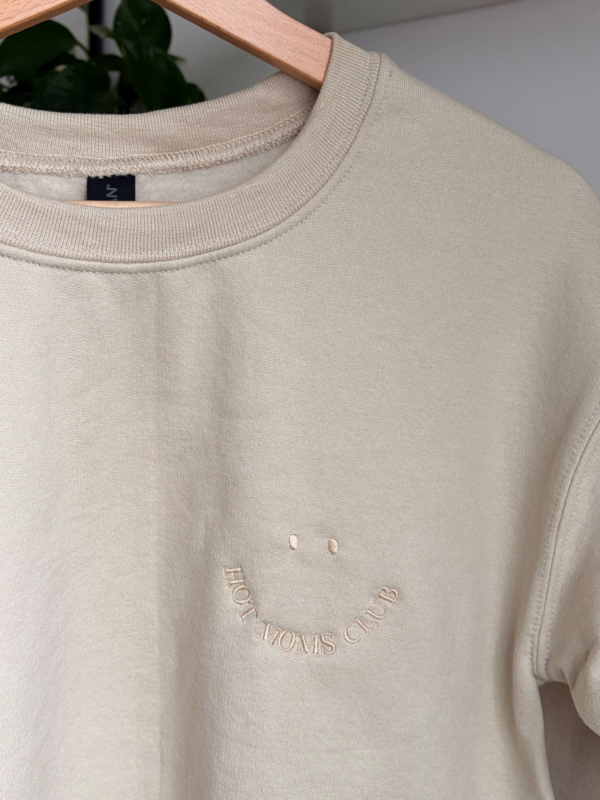 Ein beiger Sweater ist mit einem beigen Smiley auf der Brust bestickt: Hot Moms Club.
