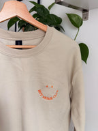 Ein beiger Sweater ist mit einem orangenen Smiley auf der Brust bestickt: Hot Moms Club.
