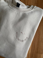 Ein gefaltetes beiges Sweatshirt liegt auf einem Holztisch. Auf der Brust ist ein lächelndes Gesicht aufgestickt, wobei der Mund aus dem Schriftzug "COOL UNCLES CLUB" in beiger Farbe besteht und zwei Punkte darüber die Augen darstellen.