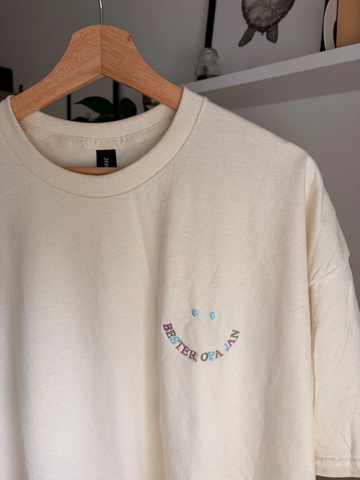 Ein T-Shirt in der Farbe Beige mit einer bunten Stickerei vorne links auf der Brust in Form eines Smileys: Bester Opa Jan.