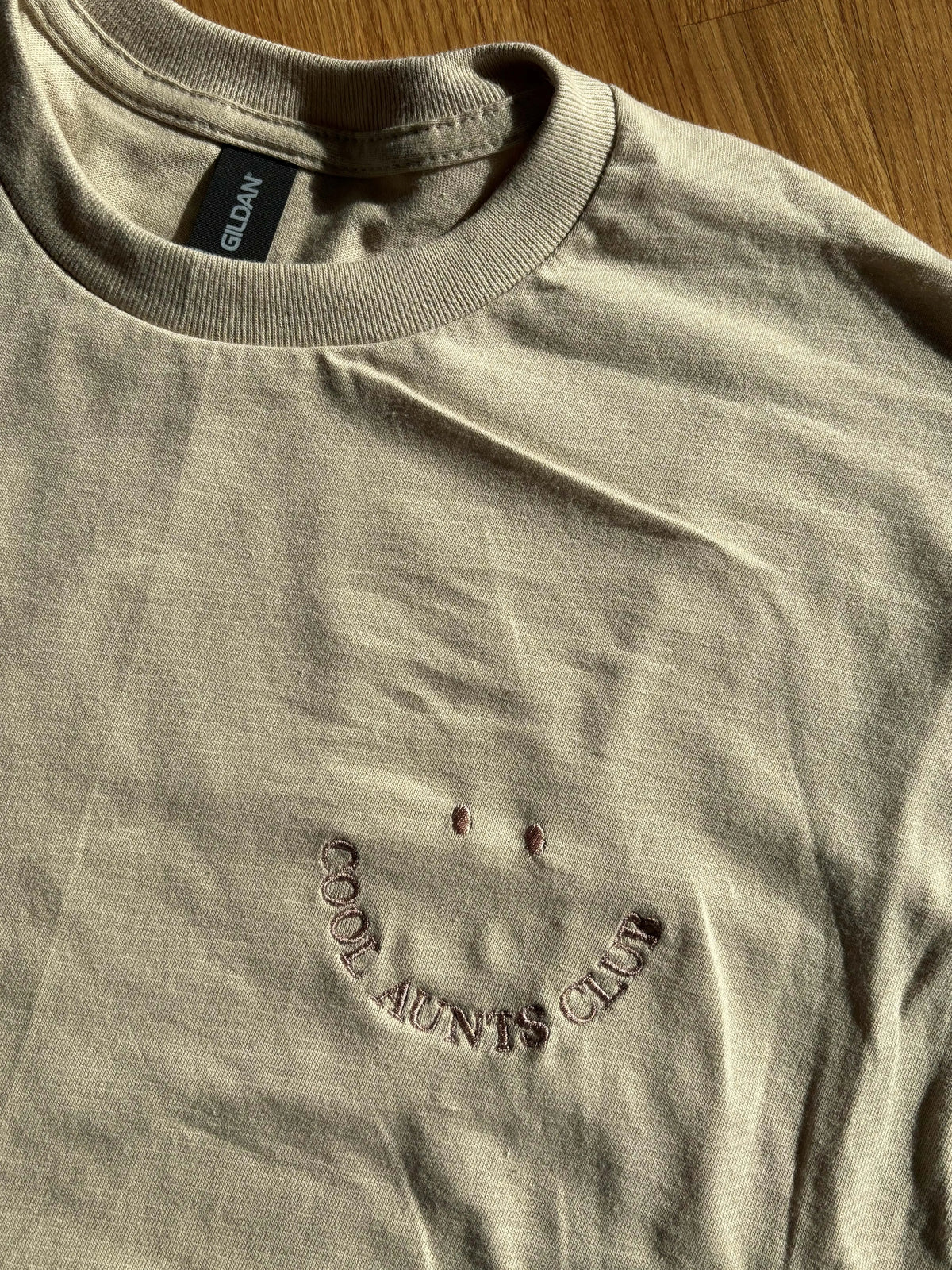 Beiges T-Shirt mit Rundhalsausschnitt. Das Shirt hat ein gesticktes Smiley-Gesicht und die Worte "COOL AUNTS CLUB" in einem geschwungenen Design. Der Text und das Smiley-Gesicht sind in verschiedenen Farben.