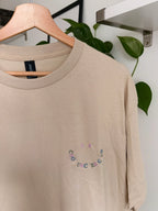 Ein beiges kurzärmeliges T-Shirt mit einem kleinen gestickten Smiley-Gesicht auf der linken Brust. Der Mund des Smiley-Gesichts wird durch den mehrfarbigen, bogenförmigen Schriftzug "COOL UNCLES CLUB" gebildet, ergänzt durch zwei lila Punkte.