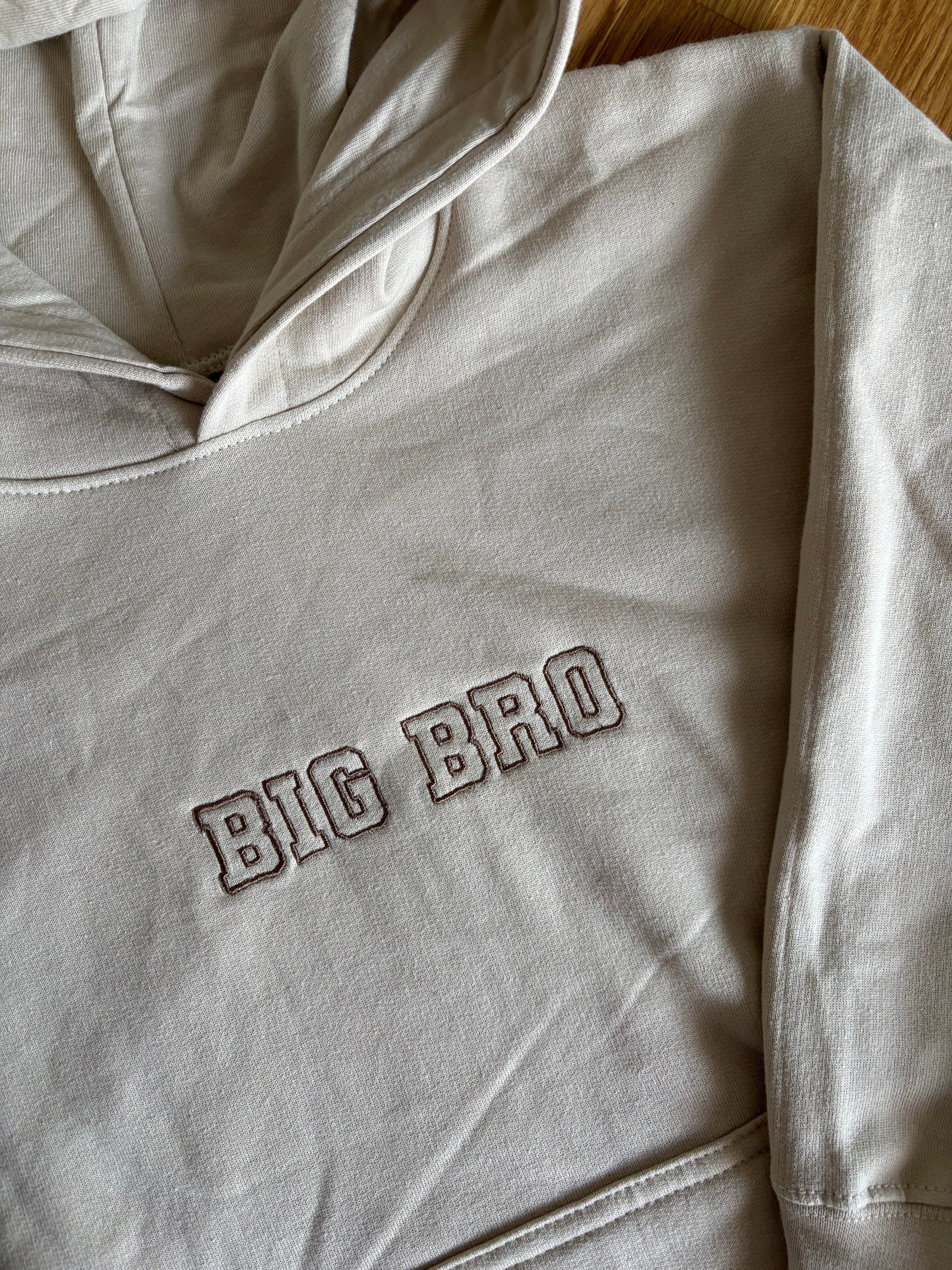 Ein beiger Pullover mit Kapuze mit einem Stick vorne BIG BRO in braun.