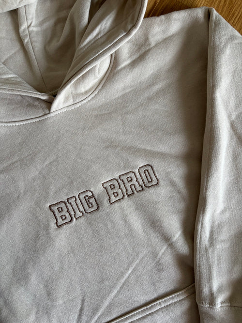 Ein beiger Pullover mit Kapuze mit einem Stick vorne BIG BRO in braun.