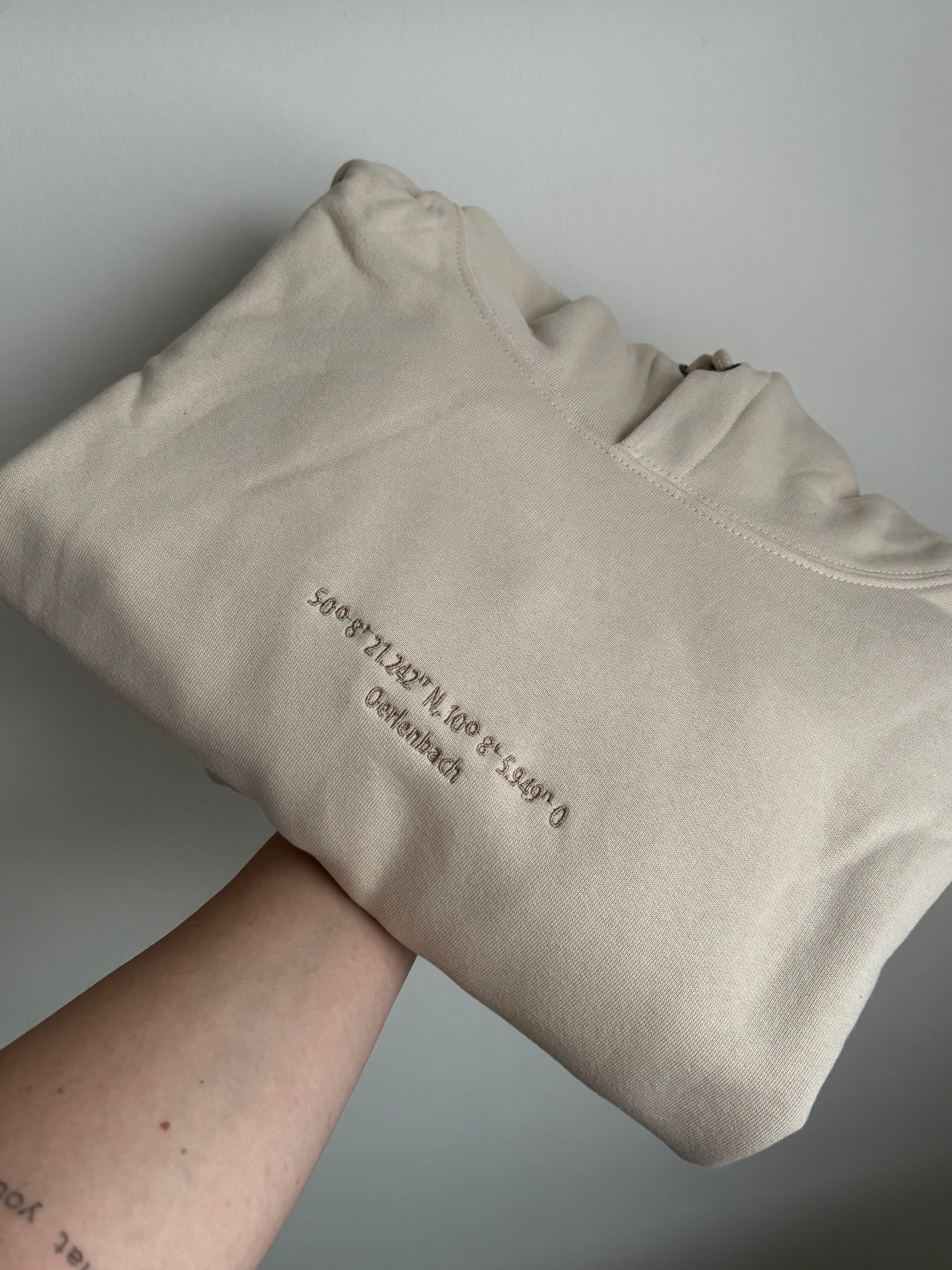 Ein beiger Pullover mit Kapuze gefaltet. Auf der Brust sind minimalistisch Koordinaten und der Name der Heimatstadt gestickt.