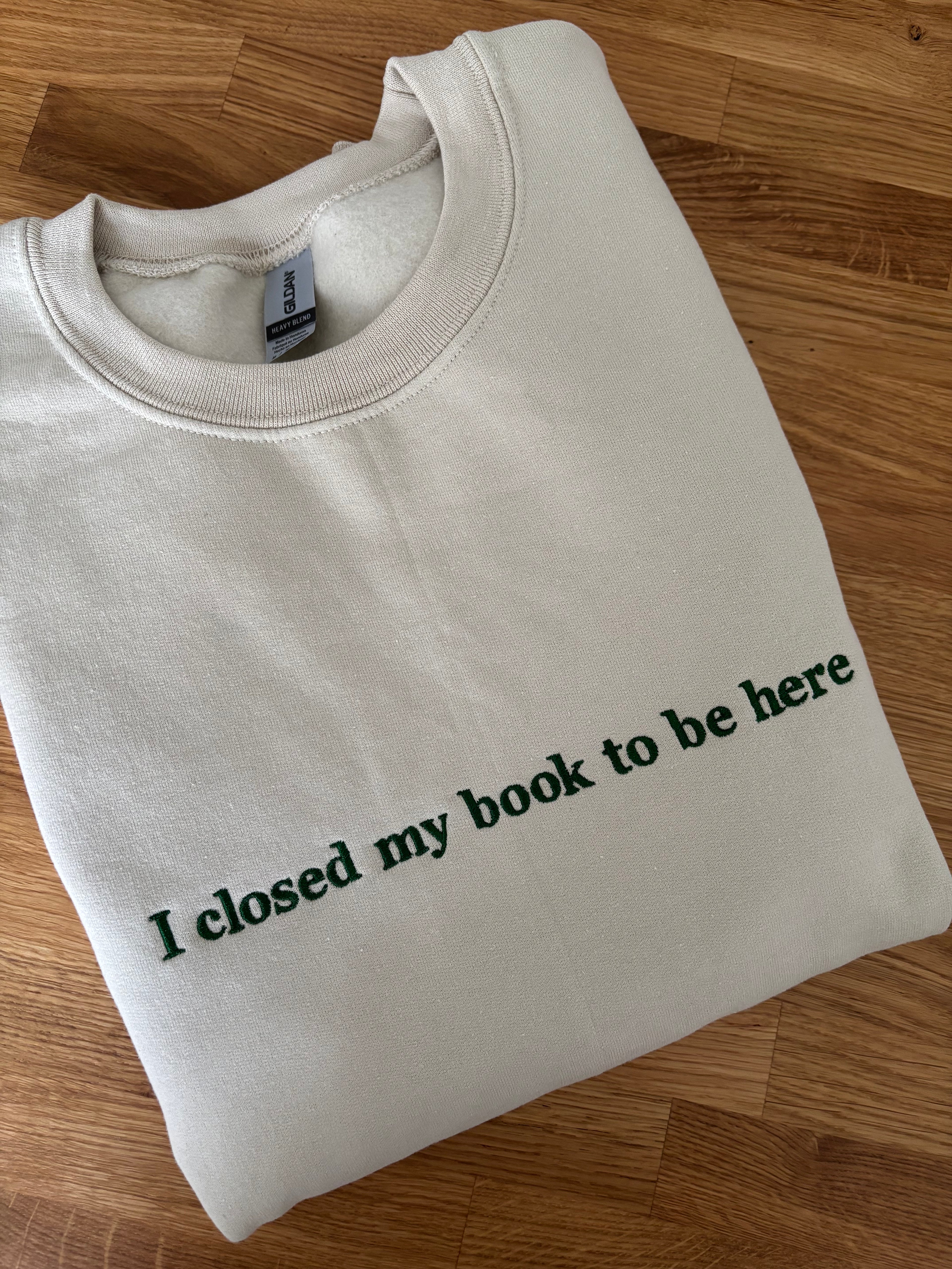 Ein beiger Sweater mit einem dunkelgrünen Stick auf der Brust "I closed my book to be here".