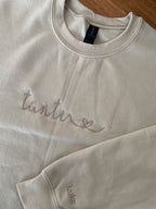 Ein beiger Sweater mit einem beigen Stick auf der Brust "Tante" mit einem Herz. Auf dem Ärmel ist der Name "Luke" gestickt.