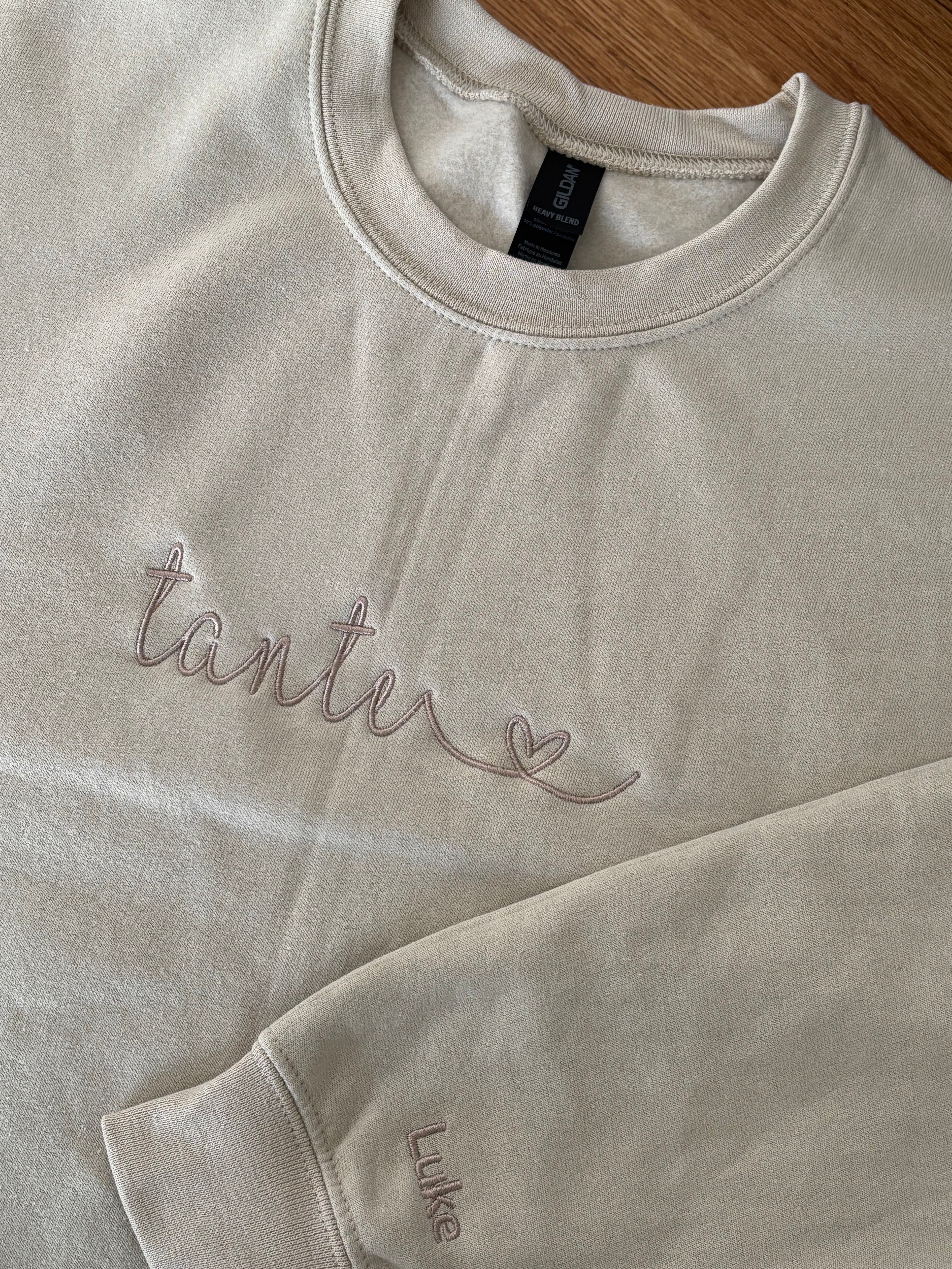 Ein beiger Sweater mit einem beigen Stick auf der Brust "Tante" mit einem Herz. Auf dem Ärmel ist der Name "Luke" gestickt.