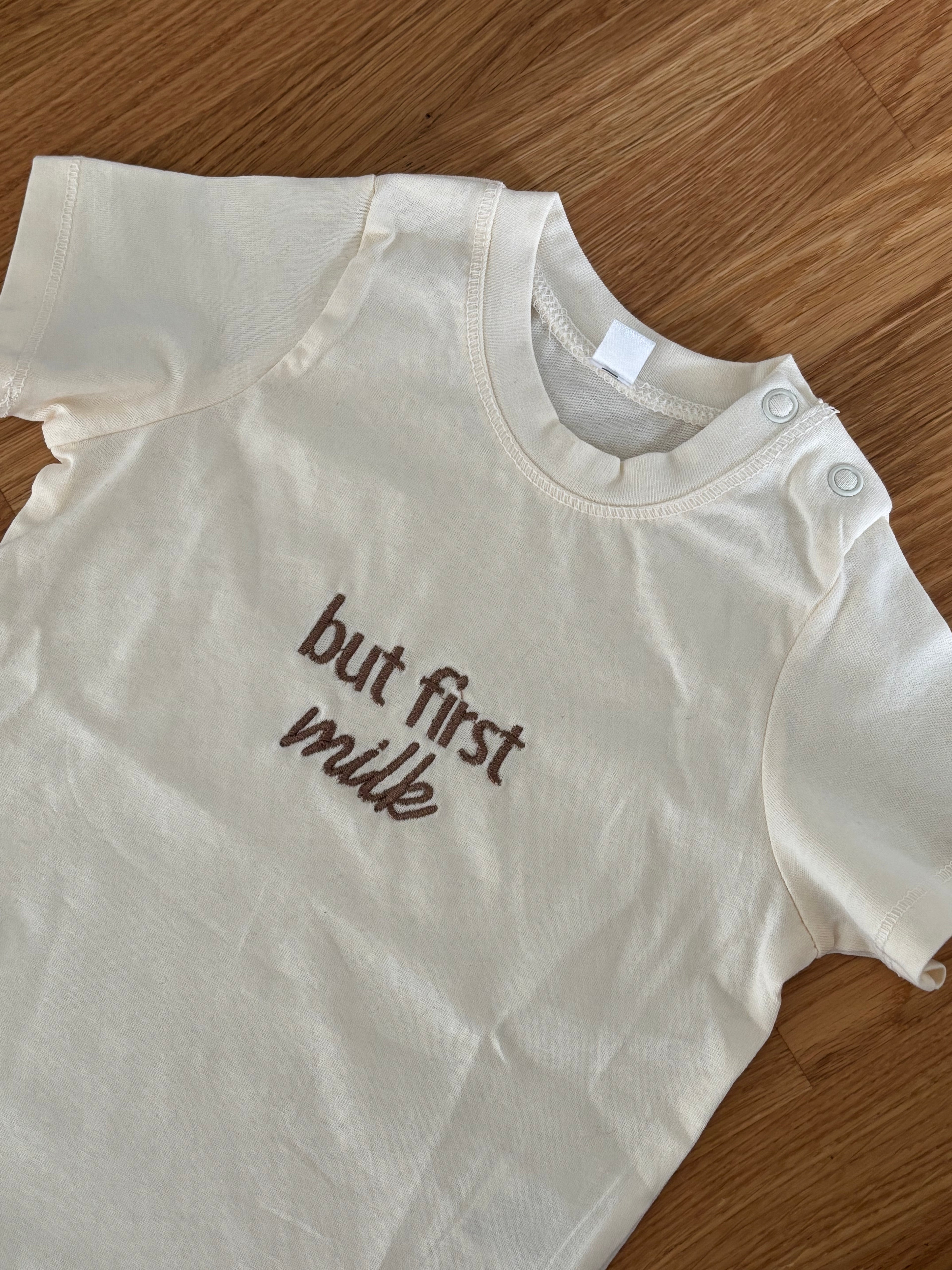 Ein beiges Kinder T-Shirt mit zwei Knöpfen an der Schulter. Vorne mittig ist in braun gestickt "but first milk".