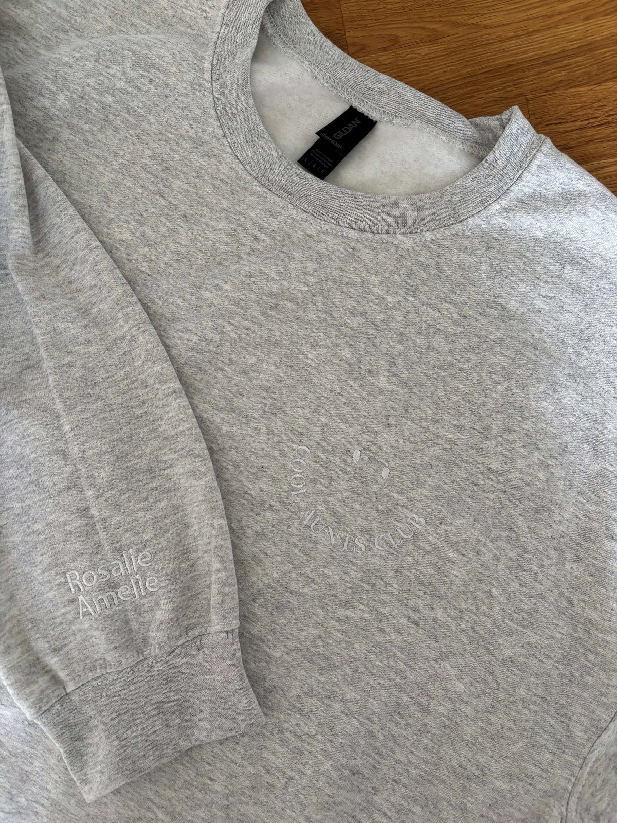 Ein Sweater in der Farbe Ash Heather mit einer Stickerei in Form eines Smileys "Cool Aunts Club" mit den Kindernamen auf dem Ärmel. 