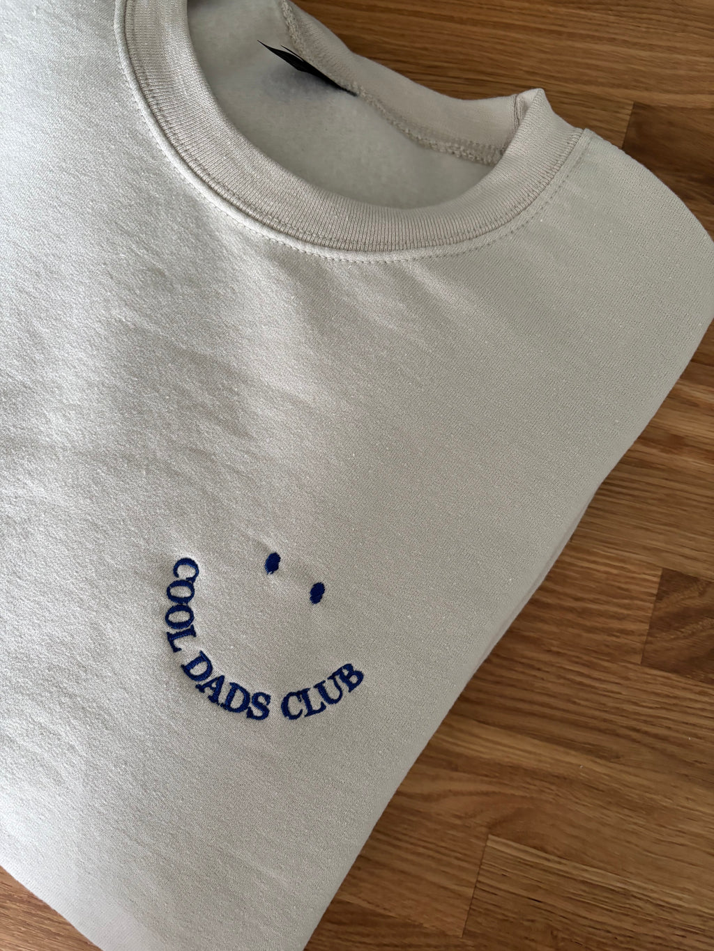 Ein gefaltetes beiges Sweatshirt liegt auf einem Holztisch. Auf der Brust ist ein lächelndes Gesicht aufgestickt, wobei der Mund aus dem Schriftzug "COOL DADS CLUB" in dunkelblauer Farbe besteht und zwei Punkte darüber die Augen darstellen.
