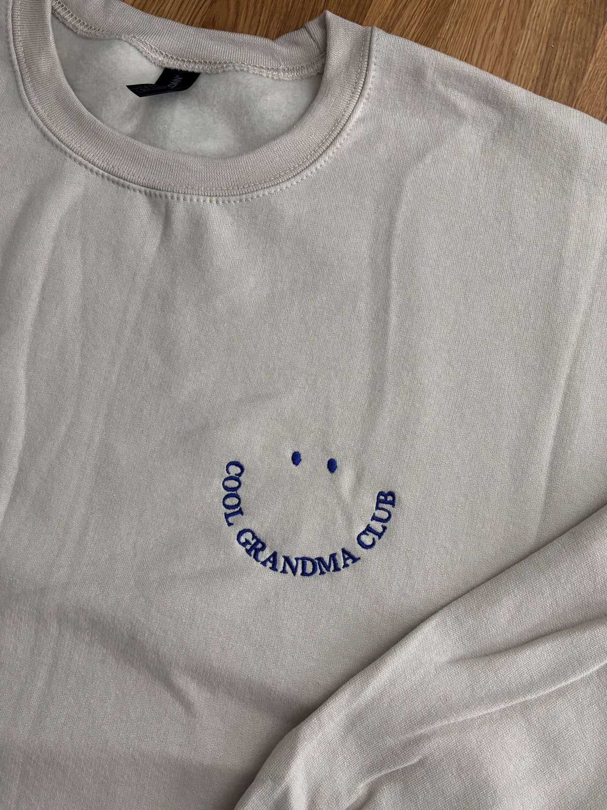 Beiges Sweatshirt mit blau gesticktem Schriftzug "COOL GRANDMA CLUB", der ein lächelndes Gesicht bildet. Das langärmlige Oberteil hat einen Rundhalsausschnitt und eine lockere Passform für den Alltag.
