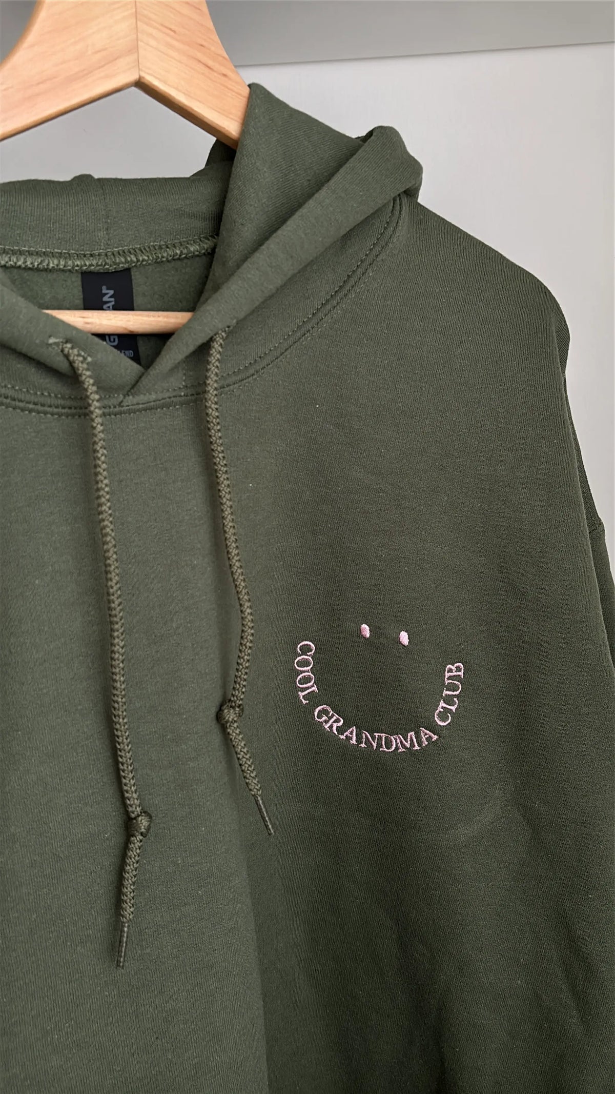 Grüner Hoodie mit lila gesticktem Schriftzug "COOL GRANDMA CLUB", der ein lächelndes Gesicht bildet. Das langärmlige Oberteil hat einer Kapuze und eine lockere Passform für den Alltag.