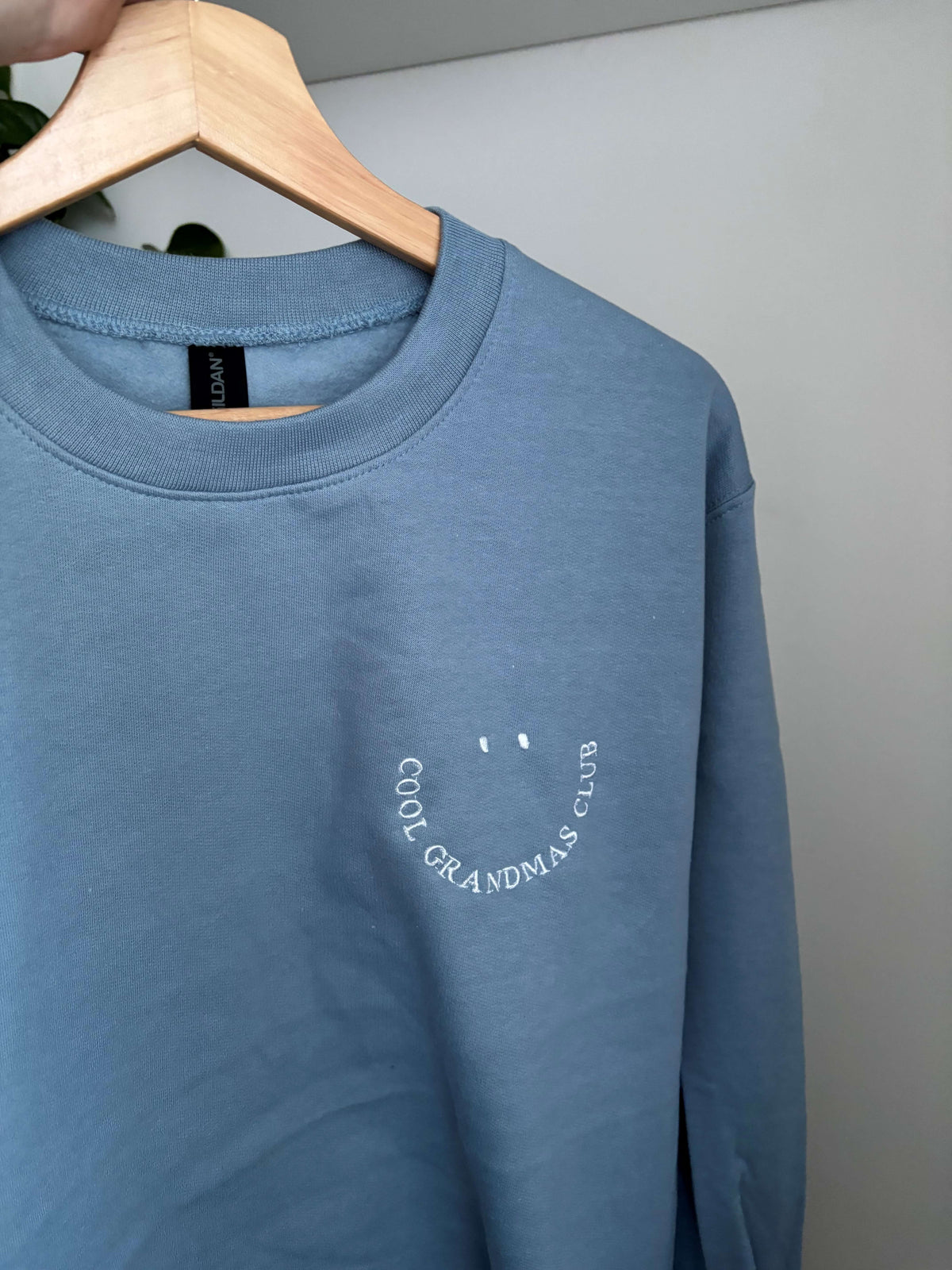 Blaues Sweatshirt mit weiß gesticktem Schriftzug "COOL GRANDMA CLUB", der ein lächelndes Gesicht bildet. Das langärmlige Oberteil hat einen Rundhalsausschnitt und eine lockere Passform für den Alltag.