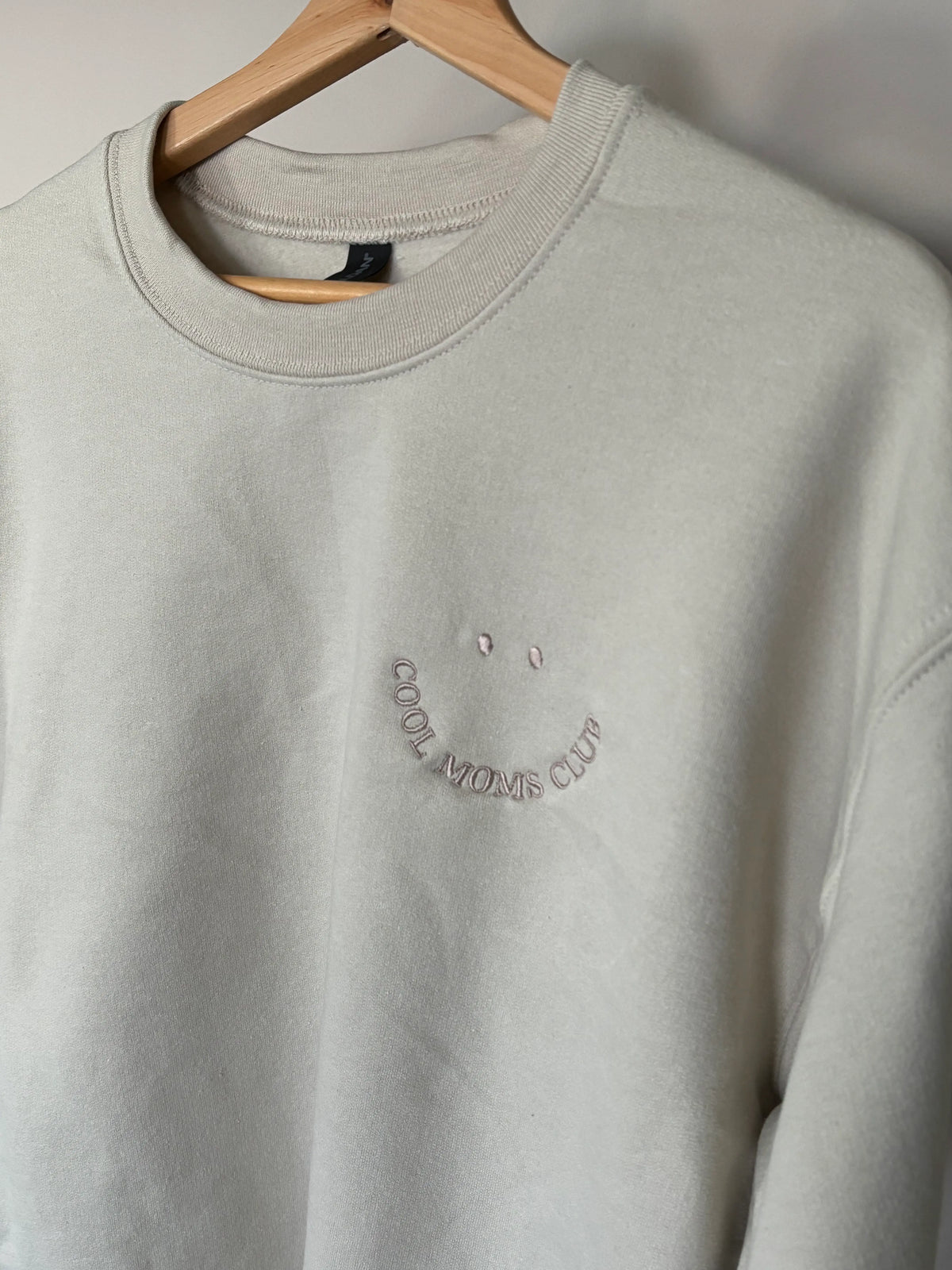 Der Cool Moms Club Sweater ist hier in beige mit beigem Garn bestickt. Der Stick sieht aus wie ein Smiley.