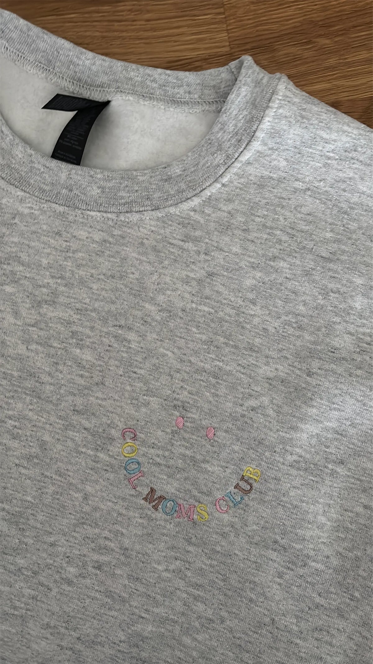 Der Cool Moms Club Sweater ist hier in Ash Heather mit buntem Garn bestickt. Der Stick sieht aus wie ein Smiley.