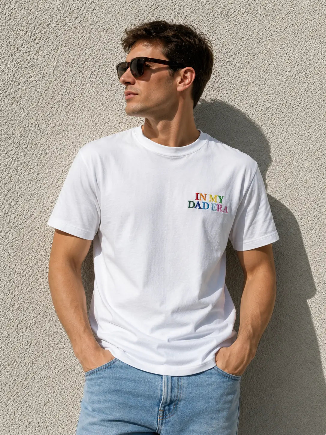 Ein Mann trägt ein weißes personalisiertes T-Shirt, welches bestickt ist. Vorne auf der Brust ist "IN MY DAD ERA" in bunten Farben gestickt. 