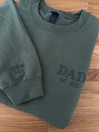 Bestickter Sweater in Military Green auf einem Holztisch. Der Sweater ist mit Dad est 2020 bestickt. Auf den Ärmel sind zwei Kindernamen zu lesen.