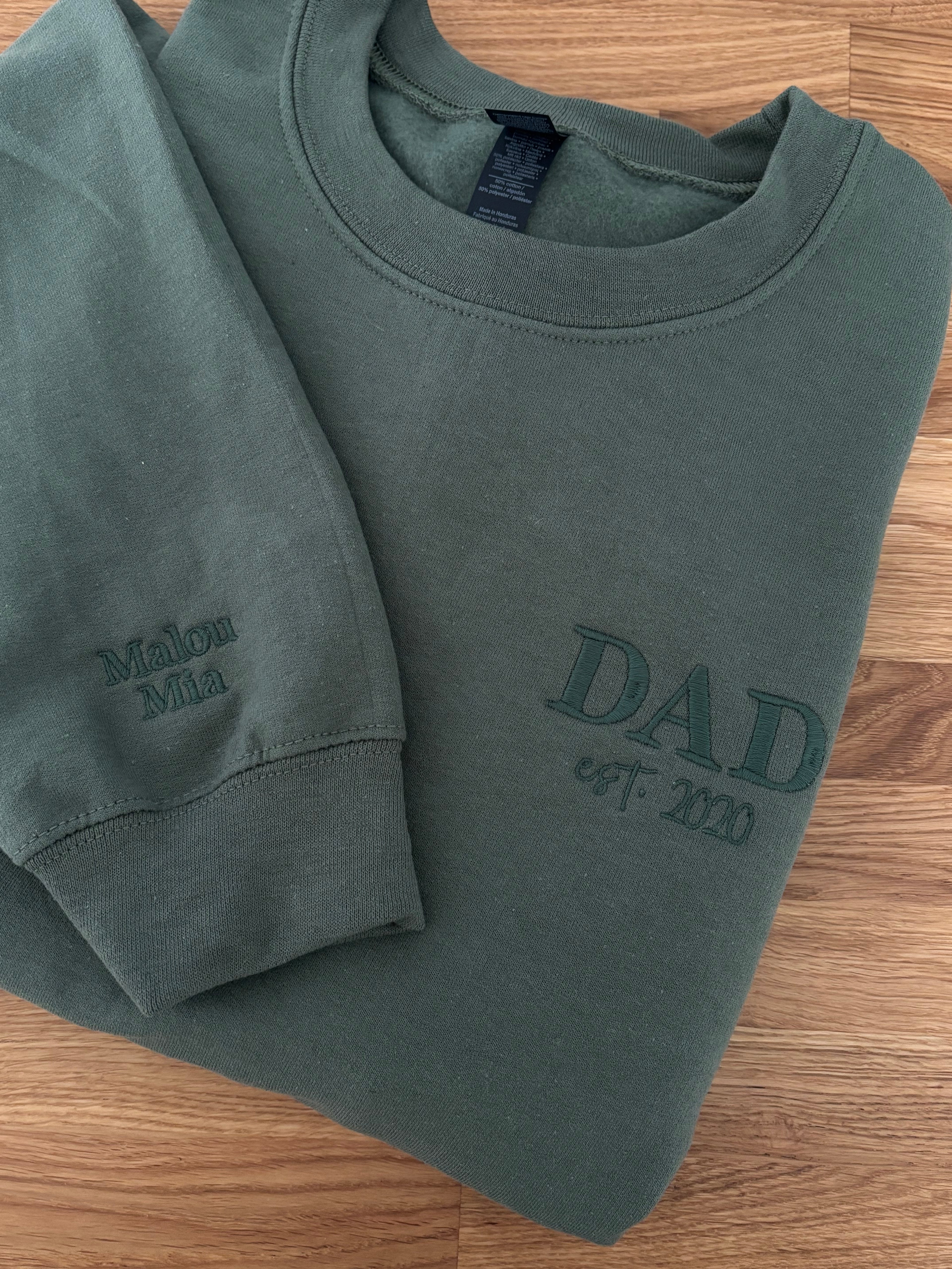 Bestickter Sweater in Military Green auf einem Holztisch. Der Sweater ist mit Dad est 2020 bestickt. Auf den Ärmel sind zwei Kindernamen zu lesen.