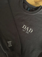 Ein dunkelbrauner Sweater. Der Stick ist in dunkelbeige. Gestickt ist in Großbuchstaben DAD und darunter est. 2018. Auf dem Ärmel stehen vier Kindernamen: Malea, Paul, Levi und Noah.