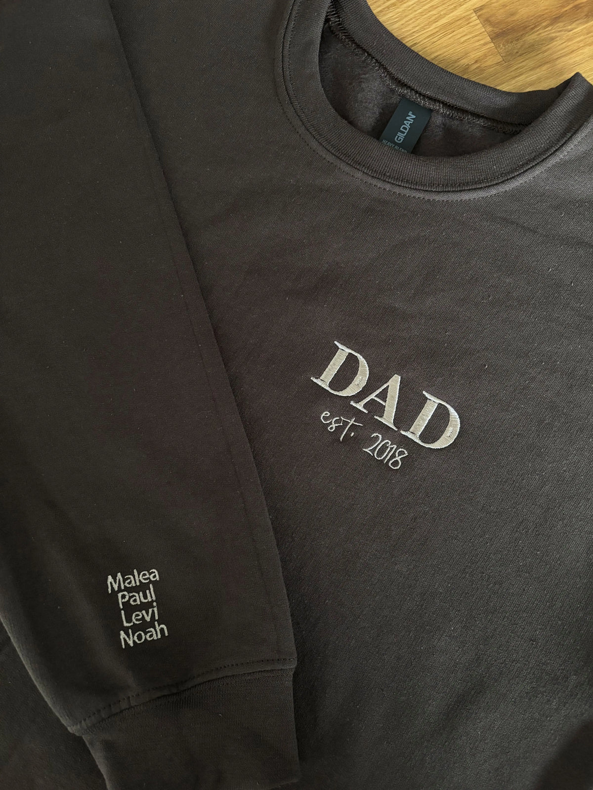 Ein dunkelbrauner Sweater. Der Stick ist in dunkelbeige. Gestickt ist in Großbuchstaben DAD und darunter est. 2018. Auf dem Ärmel stehen vier Kindernamen: Malea, Paul, Levi und Noah.
