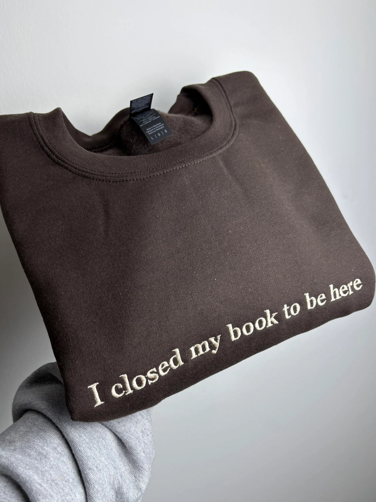 Ein Sweater in der Farbe Dark Chocolate mit dem Schriftzug I closed my Book To be Here. Der Sweater ist mit einem beigen Garn bestickt.