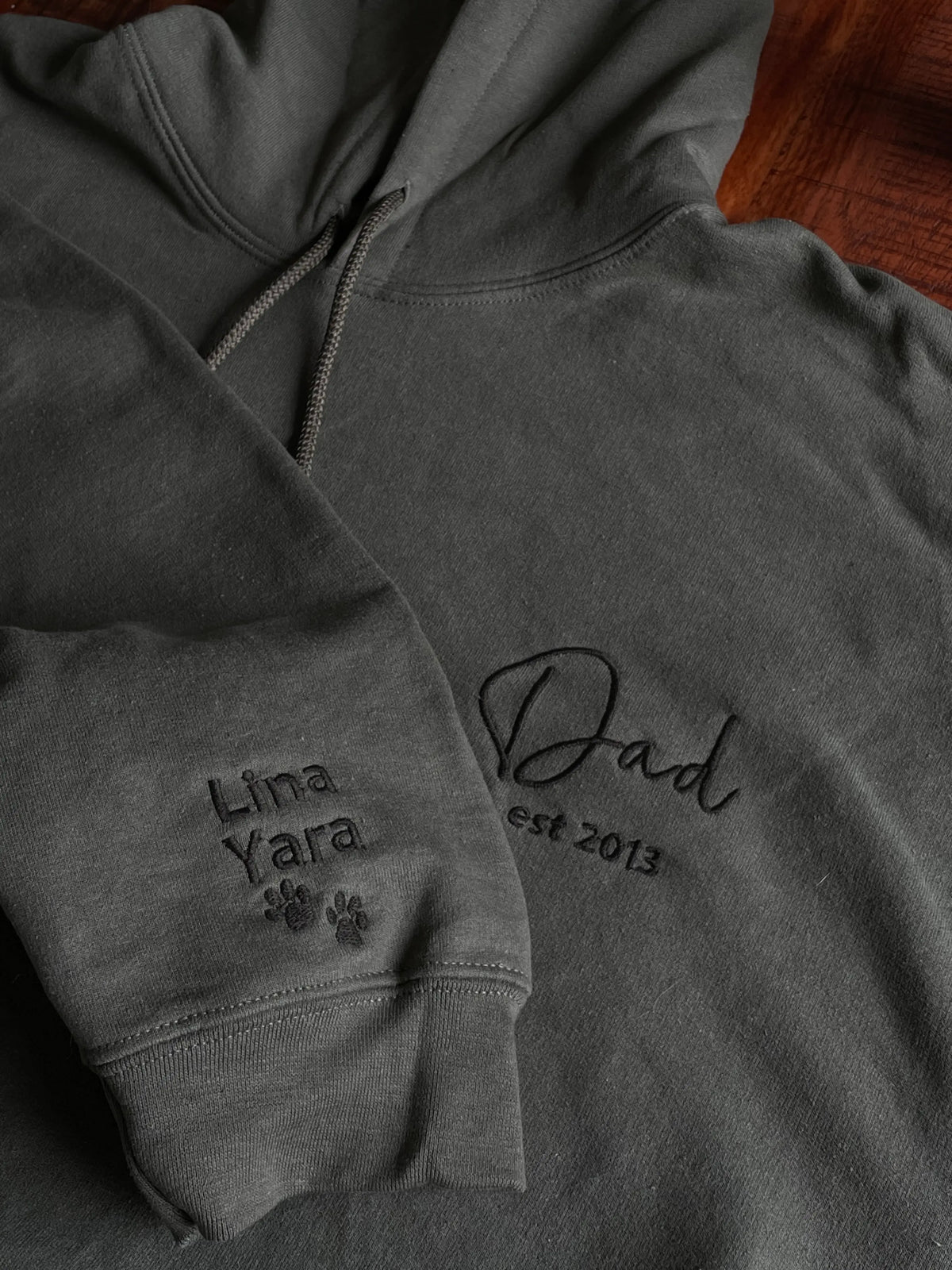 Ein Hoodie in der Farbe Dark Grey mit einer Stickerei in der Farbe Schwarz: Dad est. 2013. Auf dem rechten Ärmel sind zwei Kindernamen und zwei Pfoten gestickt: Lina und Yara.
