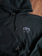 Ein Hoodie in der Farbe Forest Green mit einer Stickerei in der Farbe weiß: Opi est. 2023. Auf dem rechten Ärmel sind zwei Kindernamen gestickt: Paula und Jul. 