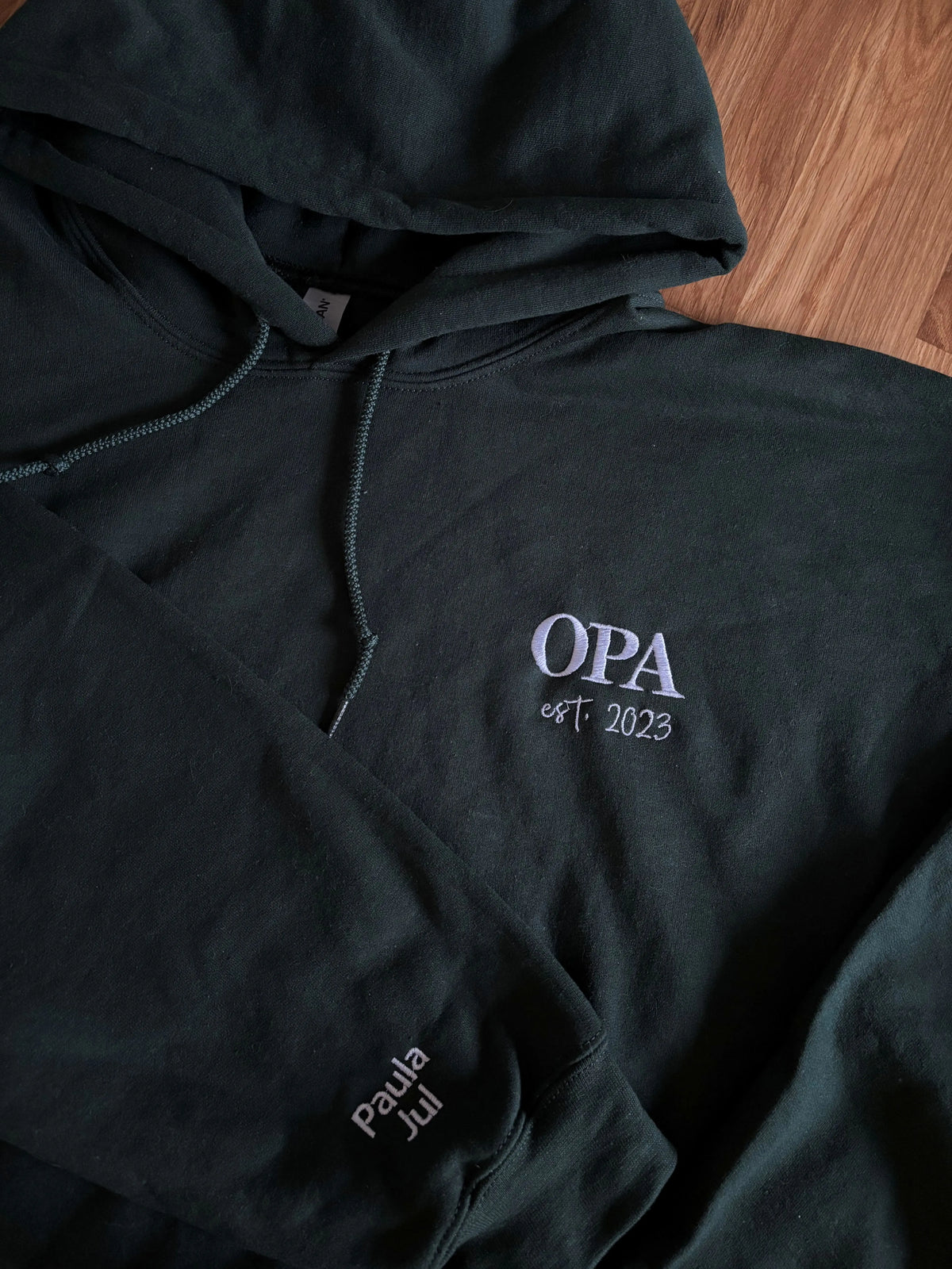 Ein Hoodie in der Farbe Forest Green mit einer Stickerei in der Farbe weiß: Opi est. 2023. Auf dem rechten Ärmel sind zwei Kindernamen gestickt: Paula und Jul. 