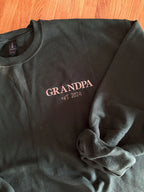 Ein Sweater in der Farbe Forest Green mit einer Stickerei in der Farbe beige: Grandpa est. 2024. 
