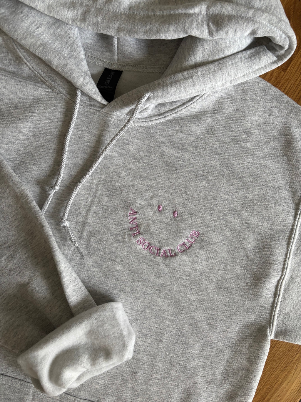 Ein bestickter Hoodie in der Farbe Ash Heather mit einem Smiley "Anti Social Club" in lila.