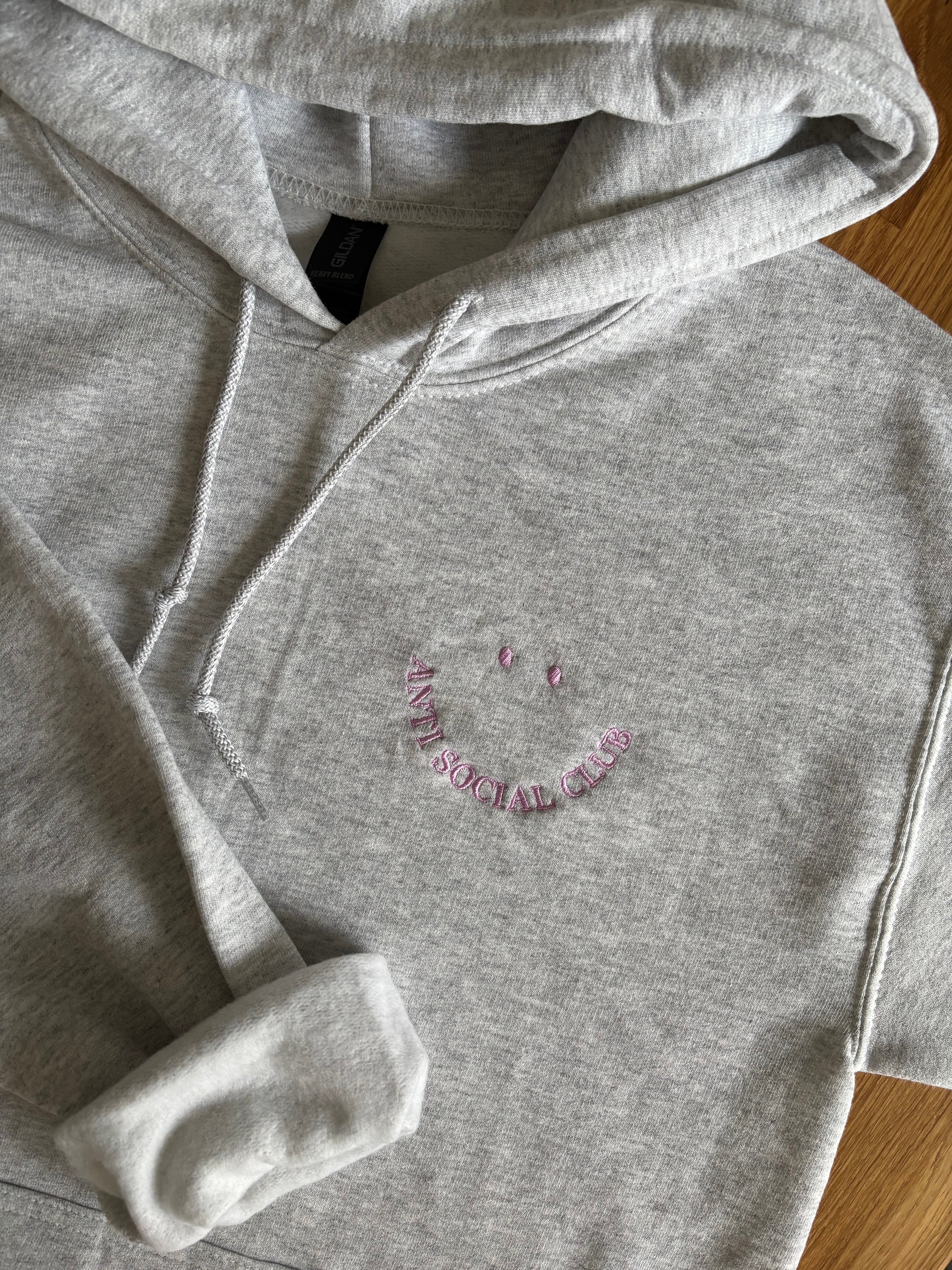 Ein bestickter Hoodie in der Farbe Ash Heather mit einem Smiley "Anti Social Club" in lila.