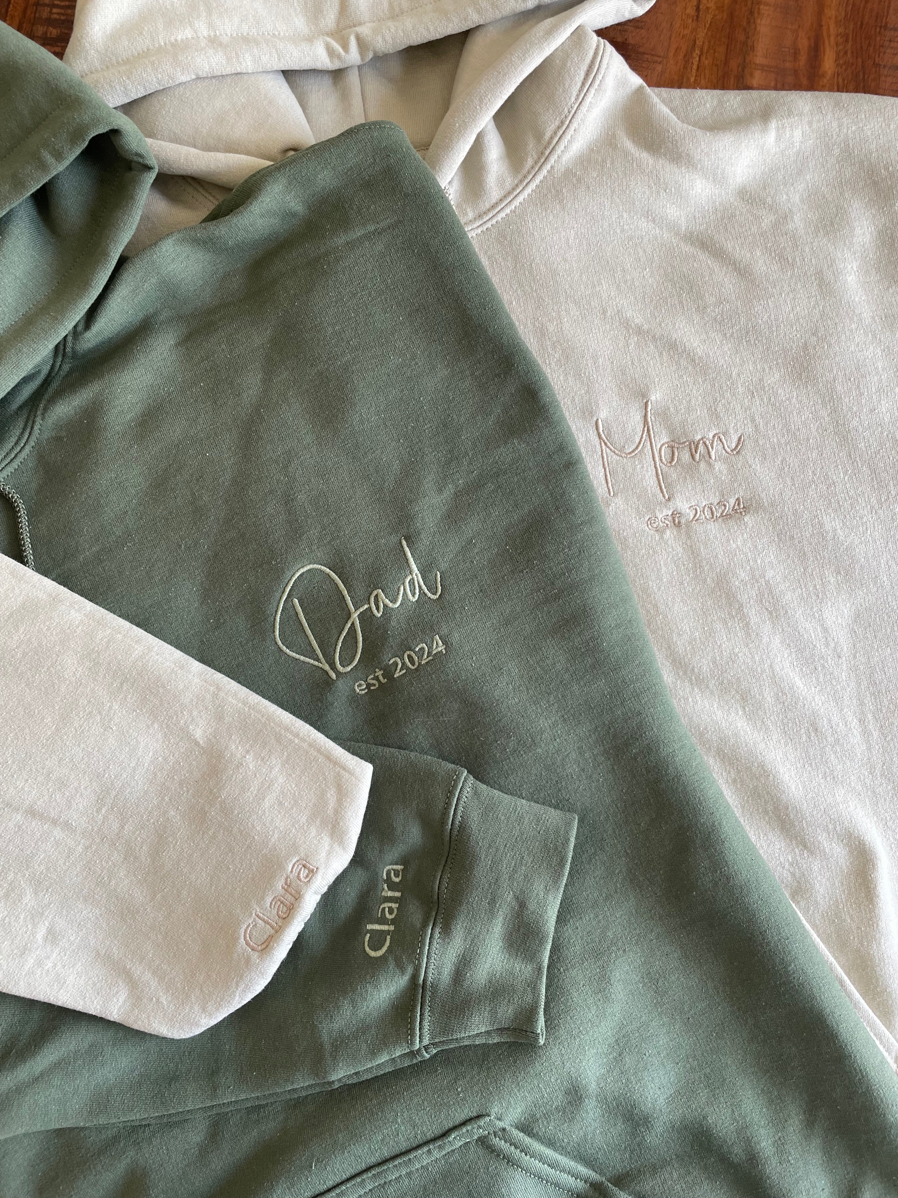 Zwei personalisierte und bestickte Hoodies mit Kapuze, worauf Mom und Dad stehen. Außerdem ist noch das Jahr, in dem sie Eltern geworden sind, gestickt. Am Ärmel ist der Name der gemeinsamen Tochter gestickt.