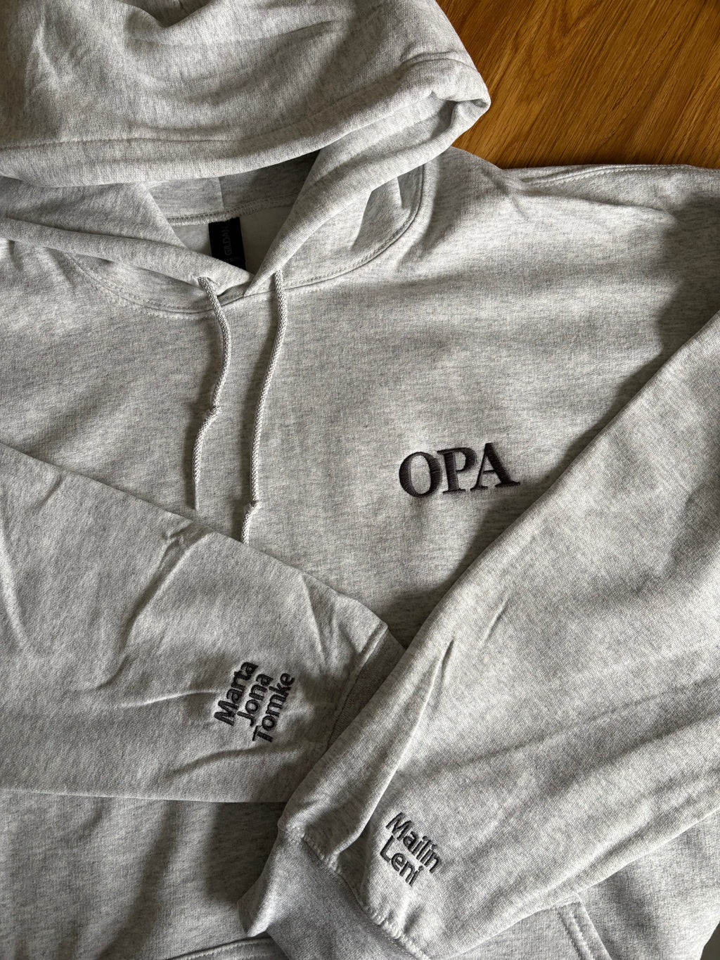Ein hellgrauer Hoodie mit einem dunkelgrauen Stick "Opa" mit den jeweiligen Kindernamen auf den Ärmeln.
